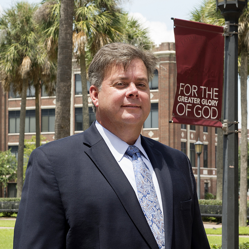 Ronald Serpas standing in front of flag on campus